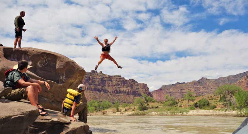 A person is suspended mid-air after jumping off a cliff into a river. Others look on, and everyone is wearing life jackets. Behind them all are tall, red canyon walls. 